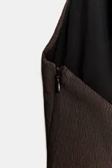 Vestido mini de color marrón oscuro con diseño de cuello halter, espalda descubierta y detalle de pliegues laterales. Presenta un bajo con encaje y abertura, cierre lateral con cremallera oculta y forro interior.