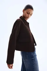 Campera corta de color marrón oscuro con cuello camisero, cierre frontal mediante botones y dos bolsillos delanteros con solapa. Presenta un diseño forrado y corte recto.