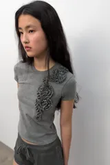 Camiseta de algodón con efecto lavado, cuello redondo y manga corta. Presenta un detalle decorativo de flores en relieve confeccionadas en el mismo tejido sobre el lado izquierdo del pecho.