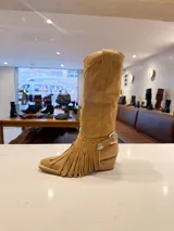 Bota de caña alta estilo western confeccionada en cuero vacuno color suela. Presenta un diseño versátil con estribo desmontable y flecos decorativos en la zona del tobillo, taco de madera y punta fina.