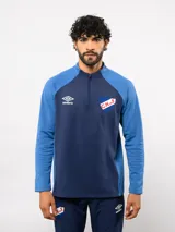Buzo de entrenamiento Umbro del Club Nacional de Football con medio cierre frontal. Presenta un diseño en bloques de color azul marino y azul brillante, con el logo de la marca y el escudo del club en el pecho.