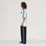 Camiseta Adidas Originals x Oasis de manga corta, color blanco con detalles en azul marino. Presenta cuello polo, las tres tiras características de la marca en los hombros, el logo del trifolio y un estampado frontal con la inscripción "oasis".