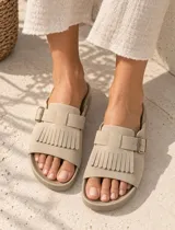 Sandalias de gamuza color marrón, con diseño de una sola tira ancha decorada con flecos y una hebilla metálica lateral ajustable. Cuenta con plantilla anatómica.
