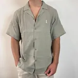 Camisa de hombre color verde oliva, de muselina con textura arrugada, manga corta, corte oversize, cuello cubano y cierre con botones. Presenta un pequeño logo bordado en el pecho.