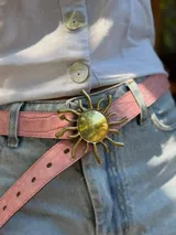 Cinturón fino rosado con hebilla metálica dorada en forma de sol.