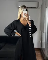 Vestido largo color marrón, con escote en V, mangas largas con puños y detalle de encaje en el pecho. Presenta un diseño asimétrico con volados en el ruedo y una abertura frontal.