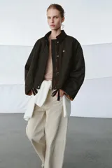 Chaqueta encerada de corte oversize, color marrón oscuro, con cuello de pana en contraste. Presenta cierre frontal con cremallera y botones a presión ocultos bajo solapa, bolsillos delanteros y mangas largas con puños ajustables.