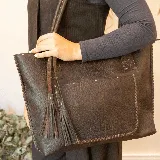 Cartera tipo tote de cuero marrón con costuras artesanales visibles, bolsillo frontal con el logo grabado, asas de hombro y detalles de flecos colgantes.