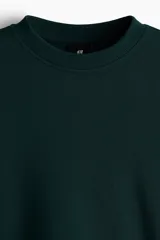 Buzo de tejido de mezcla de algodón de peso medio, color verde oscuro, con interior cepillado suave. Presenta cuello redondo ribeteado, hombros caídos y remate acanalado ancho en puños y bajo. Corte holgado.