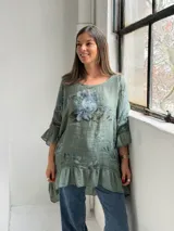 Blusa verde de lino con estampado floral en tonos azules y grises, escote redondo, mangas 3/4 con volados y ruedo asimétrico con volado fruncido.