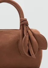 Bolso de mano tipo bowling de cuero gamuzado color marrón, con dos asas cortas y detalle de tiras con nudo y flecos colgantes en un lateral. Cierre superior con cremallera.