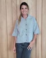 Cardigan de punto gris con cuello polo, cierre frontal con botones y mangas cortas. Presenta bolsillos frontales y terminaciones acanaladas en puños y ruedo.