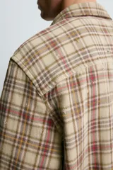 Camisa de corte cropped confeccionada en algodón, con estampado de cuadros en tonos beige, marrón y rojo. Presenta cuello solapa, cierre frontal de botones, manga larga con puño abotonado y detalles de roturas distribuidas por toda la prenda con un bajo irregular.