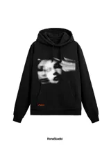 Hoodie negro de corte amplio con capucha y bolsillo frontal tipo canguro. Presenta un estampado gráfico artístico en blanco y negro en el pecho y un pequeño detalle de etiqueta naranja en la parte inferior.