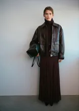 Chaqueta estilo bomber de cuero sintético en color marrón oscuro, con cuello camisero, cierre frontal de cremallera y botones a presión, bolsillos de parche con solapa y bajo elástico.