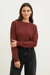 Sweater de poliéster color bordó, con cuello redondo, manga larga y diseño holgado. Presenta un patrón de punto trenzado en forma de chevrón en la parte frontal.