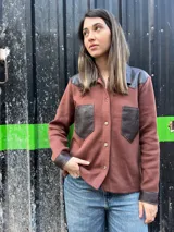 Camisa de tejido de punto color marrón con cuello, bolsillos frontales, puños y hombros de cuero sintético en color marrón oscuro, y cierre frontal con botones.