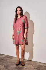 Vestido camisero corto de color rosado con estampado de aves abstractas en tonos verdes y azules. Cuenta con cuello camisero, cierre frontal con botones, mangas cortas y un bolsillo aplicado en el pecho.