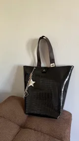 Bolso tipo tote bag negro con textura símil cuero de cocodrilo, doble asa corta marrón y correa larga al hombro. Incluye charm colgante con cadena plateada y estrella blanca.