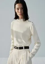 Jersey de punto fino color crema con cuello perkins y manga larga. Presenta un detalle de botones metálicos decorativos en uno de los hombros y un diseño de corte recto.