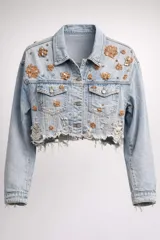 Campera de jean corta estilo cropped, en tono azul claro con efecto desgastado y ruedo desflecado. Presenta apliques decorativos de flores bordadas con lentejuelas y pedrería en tonos dorados y cobrizos distribuidos por toda la prenda.
