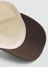 Gorro tipo gorra con visera, confeccionado en algodón, con diseño bicolor en color crudo y marrón, cierre regulable con hebilla metálica y detalle de mensaje bordado.