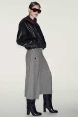 Bermuda larga de tiro alto con diseño de estampado a cuadros tipo pata de gallo. Presenta pinzas delanteras, corte amplio y un cinturón integrado que combina una tira de cuero sintético con una cadena metálica dorada. Cierre lateral con cremallera oculta.