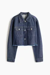 Campera cropped de denim de algodón rígido, color azul, con cuello, cierre frontal de botones y hombros caídos. Cuenta con bolsillos superiores con solapa y botón, puños abotonados y dobladillo deshilachado.