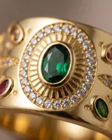 Anillo ancho de bronce con baño de oro, diseño texturizado con detalles ornamentales, piedra central verde ovalada rodeada de circonias y pequeñas piedras rojas incrustadas.