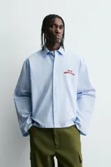 Camisa de algodón de corte regular, color celeste con finas rayas blancas verticales. Presenta un bordado en color rojo en el pecho con la frase "Café du Matin Doux".