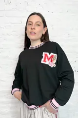 Buzo negro de rústico con cuello, puños y ruedo con ribete rosa y doble línea negra. Estampado en el pecho con la letra M en rojo y blanco, decorada con flores y un tigre.