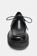 Zapato tipo oxford de cuero negro con cierre de cordones, costuras decorativas en la puntera y suela gruesa dentada de estilo track.