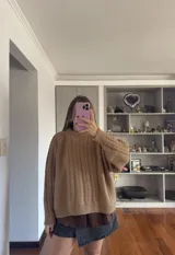 Buzo tejido de punto color tostado, con cuello redondo y diseño de trenzas verticales en el frente y mangas. Presenta un corte holgado con puños y ruedo acanalados.