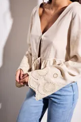Blusa color beige claro, con escote en V profundo y cierre frontal mediante lazo ajustable a la altura de la cintura. Presenta un peplum con volado y mangas largas con puños anchos con bordados calados.
