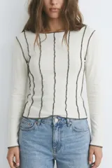 Remera de manga larga en color blanco, confeccionada en tejido de punto acanalado. Presenta un diseño ajustado con costuras externas visibles en contraste de color oscuro que recorren verticalmente la prenda.