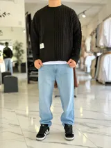 Sweater de punto oversize color marrón, con cuello redondo y diseño combinado: la mitad derecha cuenta con tejido de ochos y la izquierda es lisa. Incluye una etiqueta decorativa blanca en el frente inferior derecho.
