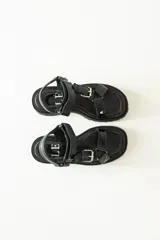 Sandalias negras de estilo chunky con plataforma dentada. Presentan tiras ajustables con cierre de velcro y hebillas metálicas, combinando texturas de cuero granulado y detalles en charol.