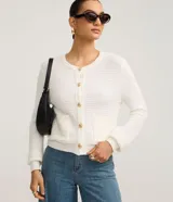 Cardigan de punto blanco, con cuello redondo, manga larga y cierre frontal de botones dorados. Posee dos bolsillos cuadrados en la parte delantera inferior.