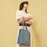 Cartera tipo tote de estructura vertical, color negro, con doble asa larga para llevar al hombro y correa desmontable y regulable. Cuenta con bolsillo frontal exterior de fácil acceso y detalles de costuras visibles.