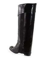 Bota alta de charol negro con caña recta, puntera redondeada y correa con hebilla en la parte superior.