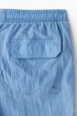 Short de baño de nylon en color azul, con cintura elástica y cordón de ajuste interno. Presenta bolsillos laterales, un bolsillo trasero con cierre autoadherente y un pequeño parche con logo en la parte inferior. Incluye suspensor interno de malla.