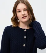 Cárdigan de punto trenzado color marrón, con cuello redondo, manga larga y cierre frontal con botones metálicos. Presenta puños y ruedo acanalados.