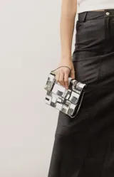 Cartera tipo sobre o clutch rectangular, cubierta de lentejuelas en tonos blanco, negro, gris y dorado formando un patrón geométrico cuadriculado. Cuenta con solapa y una fina cadena metálica.