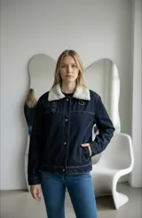 Campera de jean azul oscuro con cuello de corderito sintético color crema. Presenta cierre frontal con botones metálicos, bolsillos laterales y detalles de hebillas decorativas en la parte superior del pecho.