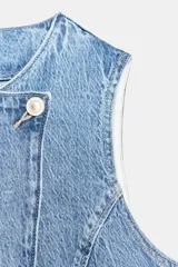 Chaleco de denim con cuello redondo y diseño sin mangas. Presenta un cierre frontal cruzado con botones y falsos bolsillos de vivo en la parte delantera.