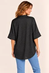 Blusa de manga corta negra con diseño texturizado en relieve de pequeños cuadrados, cuello mao y escote en V. Presenta un corte holgado con dobladillo asimétrico, siendo más larga en la parte trasera, y aberturas laterales en el bajo.