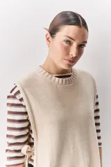 Chaleco tejido de punto color beige, con cuello redondo, terminaciones en rib y lazos laterales regulables que permiten ajustar la prenda.