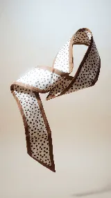 Pañuelo delgado y largo (twilly) de seda o material similar, reversible. Un lado es de color beige claro con estampado floral tropical en tonos azul marino y terracota. El otro lado es liso color beige/nude con una frase manuscrita en azul que dice "I'll finally be as you wish!".