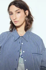 Camisa de silueta amplia y caída fluida, color celeste con rayas verticales blancas. Presenta cuello camisero clásico, cierre frontal con botones y un bolsillo aplicado en el pecho. Las costuras están resaltadas en azul oscuro.