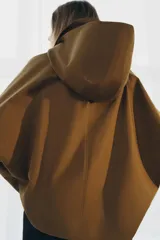 Trench corto color verde militar, con cuello solapa y capucha. Tiene manga larga con trabillas y botón en los puños, bolsillos delanteros y bajo asimétrico. Cierre frontal con botones. El tejido es repelente al agua.